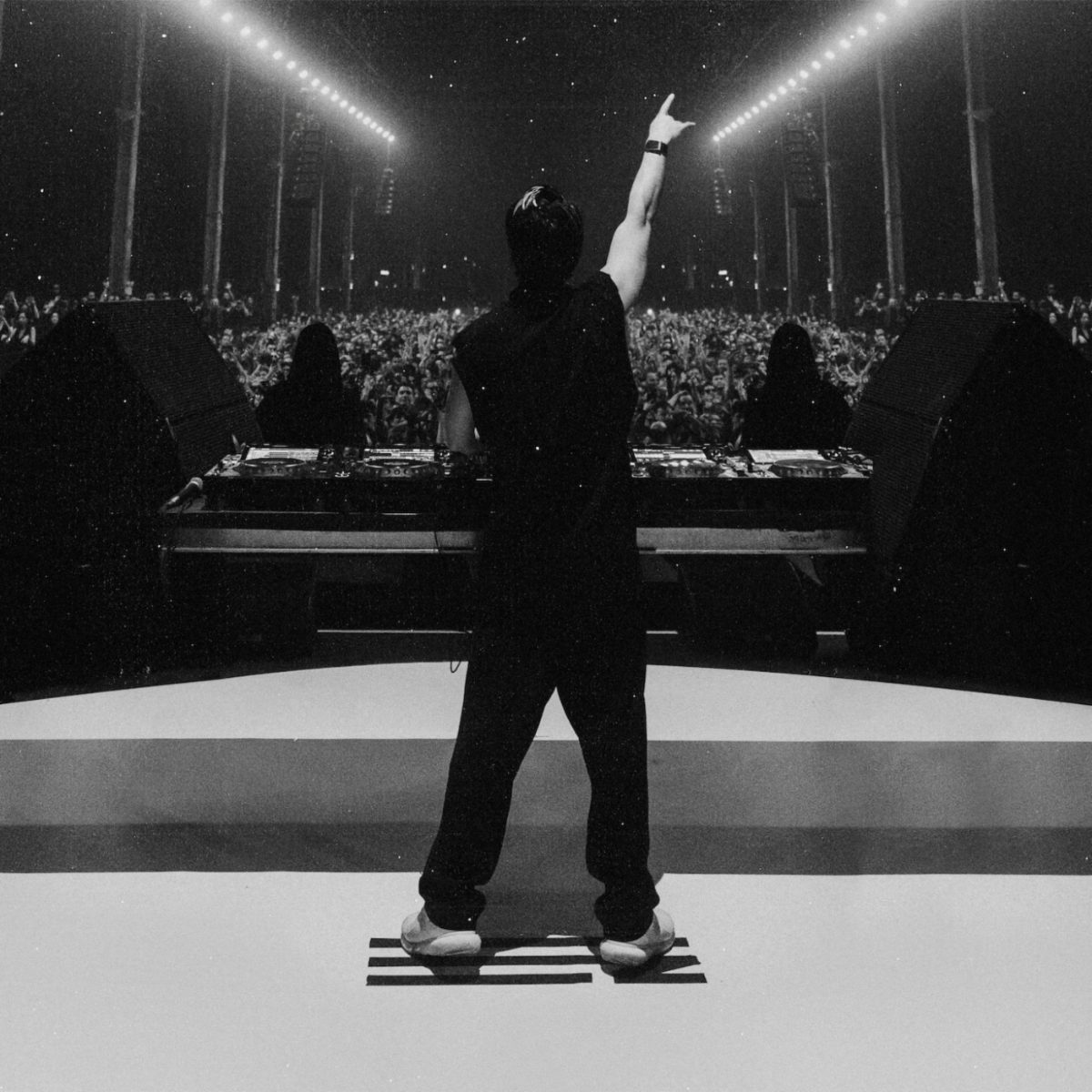 ZHU seen from behind onstage, arm raised toward a packed crowd under bright stage lights