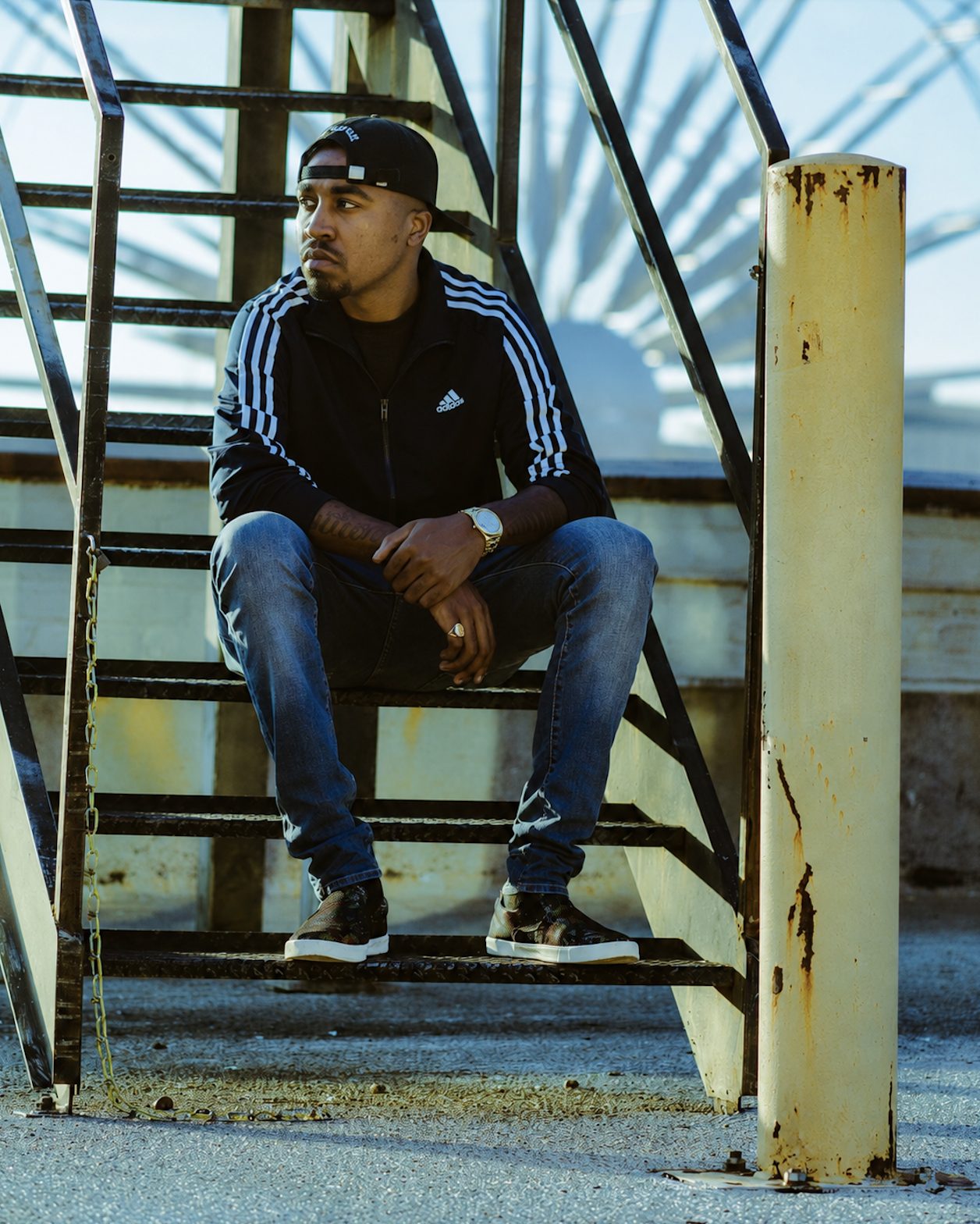 James Artissen seated on outdoor metal stairs in a backwards cap, black Adidas track jacket and jeans, gold watch visible, photographed in hard sunlight.