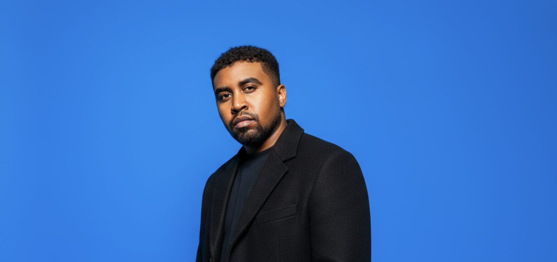 James Artissen in a black overcoat and black tee, photographed against a solid bright blue backdrop, looking directly into the camera.