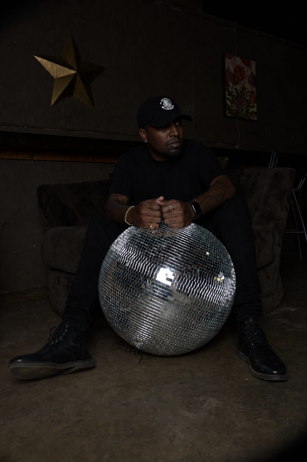 James Artissen dressed in all black, seated in a dark room with his hands resting on a large mirror ball, a gold star on the wall behind him.