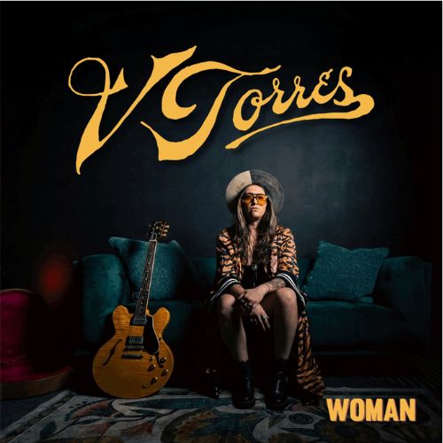 Cover artwork for V Torres’ album “Woman”: the artist seated on the floor in front of a teal velvet sofa with a hollow-body electric guitar beside her, under a gold script logo reading V Torres.