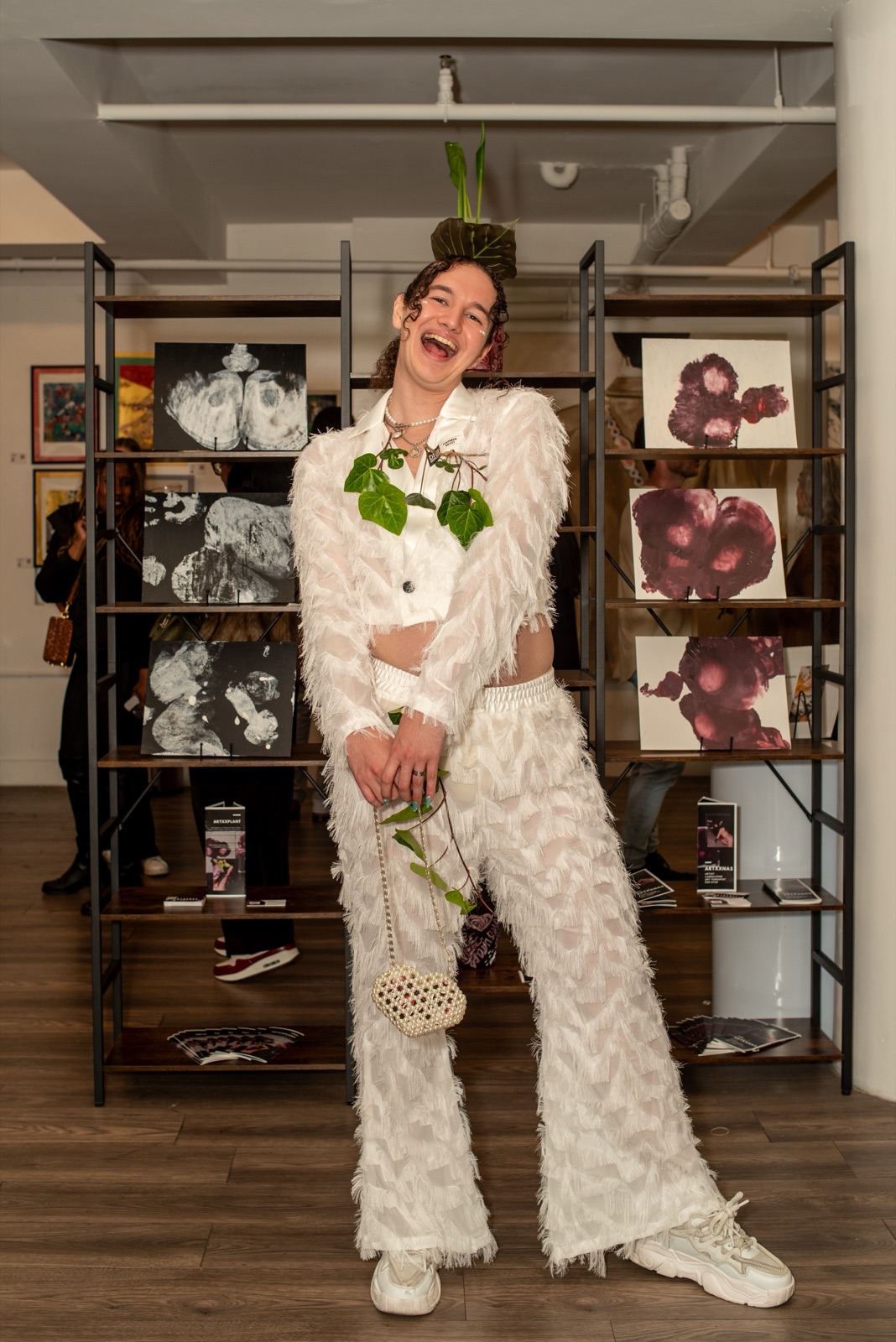 artxxnas laughing in an all-white textured suit with a plant balanced on his head, standing in front of gallery shelves displaying the burgundy and black-and-white ARTXXSEX body-print paintings.