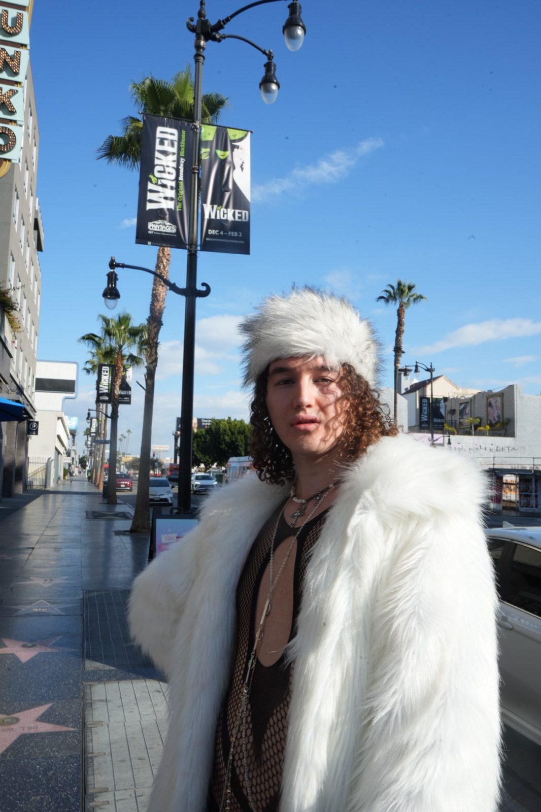 artxxnas stands on Hollywood Boulevard in a white faux-fur coat and matching fur hat over a mesh top, with palm trees, theatre banners and Walk of Fame stars behind him.