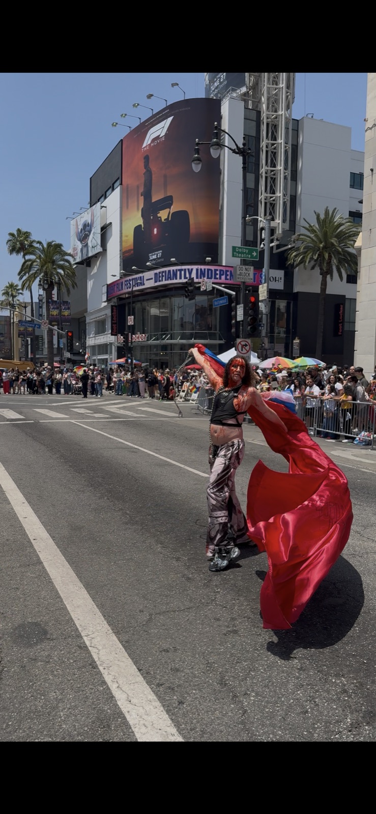 artxxnas performs in the middle of Hollywood Boulevard during LA Pride 2025, covered in simulated blood and trailing a large red fabric behind him as a crowd watches from behind barricades.