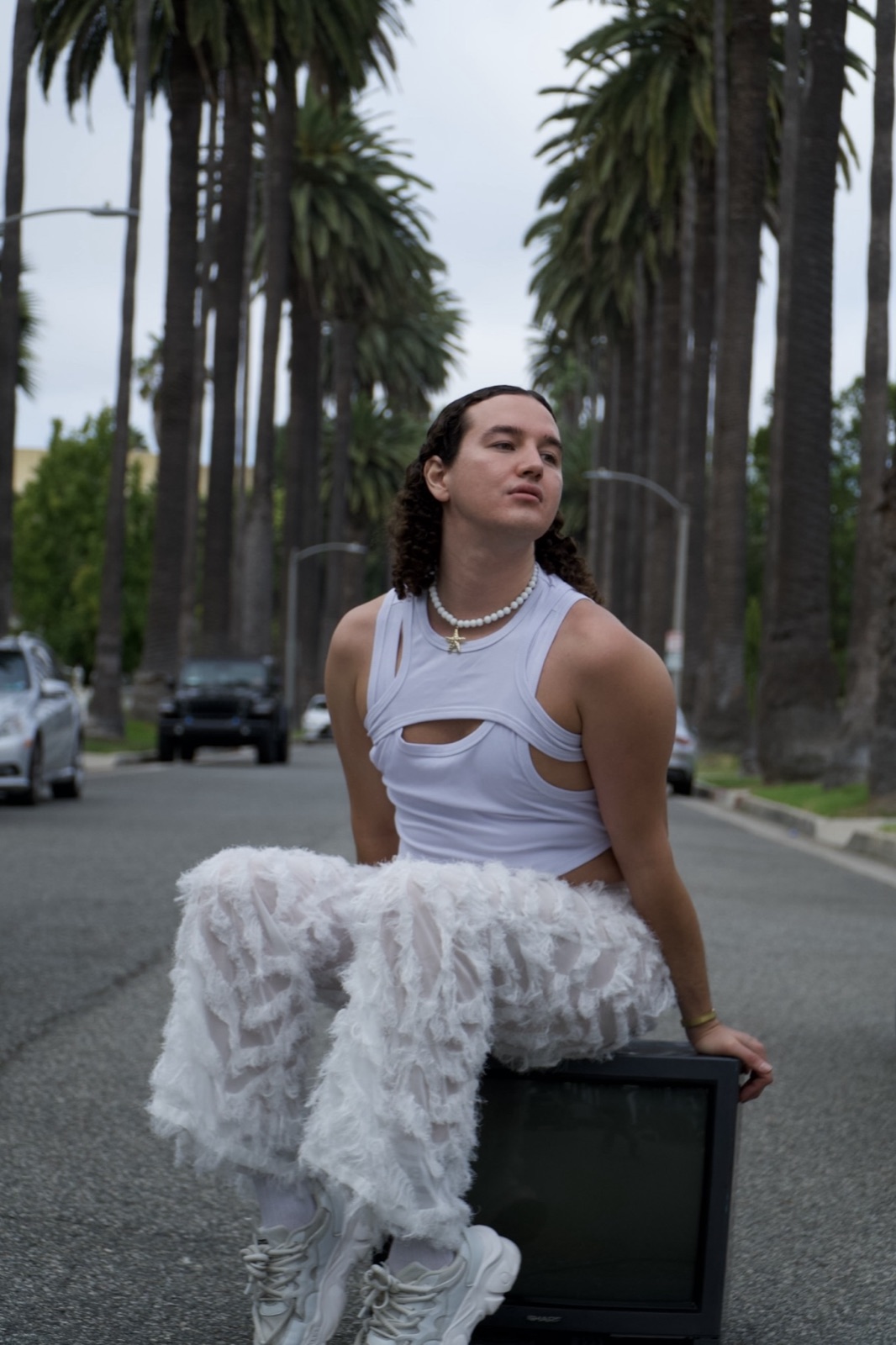 artxxnas sits on an old television set in the middle of a palm-lined Los Angeles street, wearing a cut-out white tank top, pearl necklace and layered white tulle trousers.