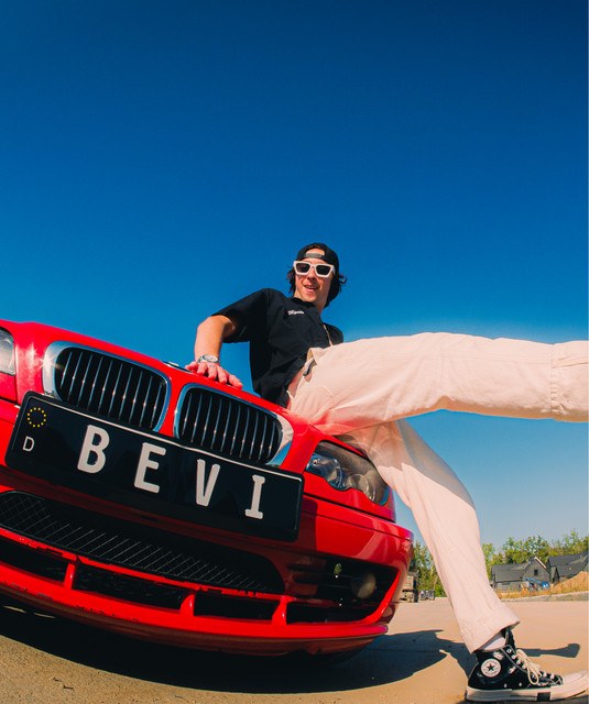 BEVIxPlanett in white sunglasses and cream trousers, one leg kicked out, leaning on the hood of a red BMW with a BEVI license plate under a clear blue sky