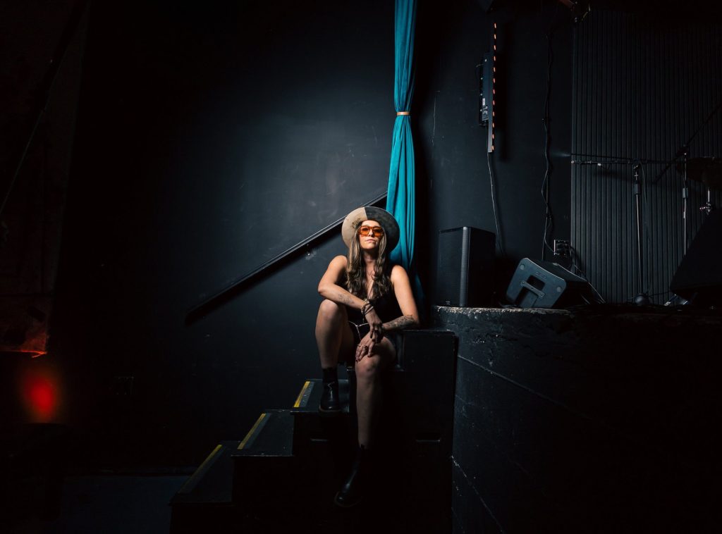 V Torres sits on the steps beside a darkened stage in a wide-brimmed hat and orange sunglasses, lit by a single spotlight.
