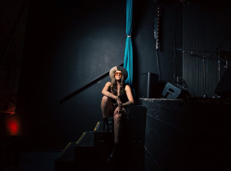 V Torres sits on the steps beside a darkened stage in a wide-brimmed hat and orange sunglasses, lit by a single spotlight.