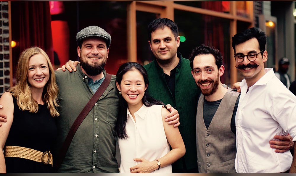 The six members of folk-classical collective Floyds Row standing arm in arm outside a venue at night, smiling at the camera.