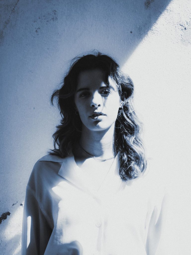 Blue-toned monochrome portrait of Italian singer-songwriter Giuliana May, standing against a sunlit wall with a diagonal shadow, wearing a pale shirt and thin chain necklace.