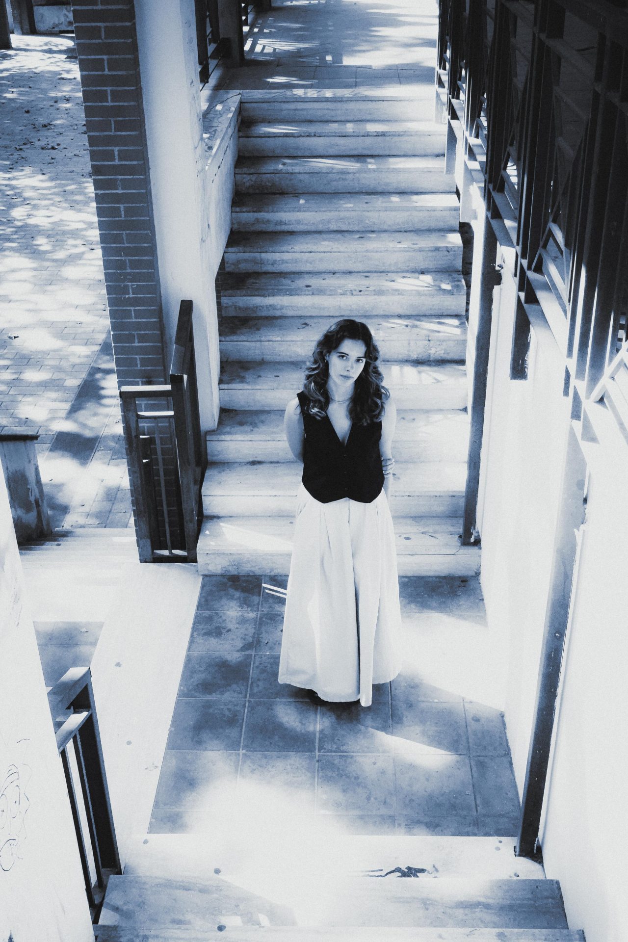 Giuliana May in a black vest and long pale skirt standing at the foot of a stark sunlit staircase, photographed from above in cold blue monochrome.