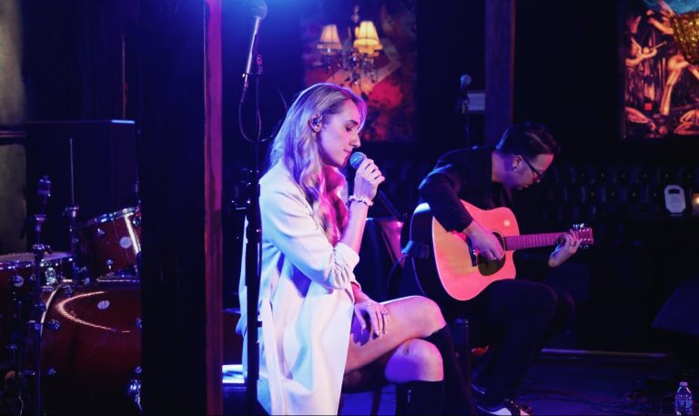Singer-songwriter Lindsay Boven seated on a stool under pink and purple stage light, singing into a handheld microphone beside an acoustic guitarist