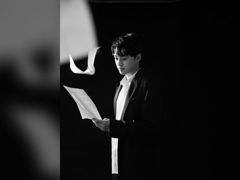 Black-and-white portrait of Los Angeles composer Yuxuan Liu in a dark suit, reading a handwritten music score as loose pages float in the air beside him