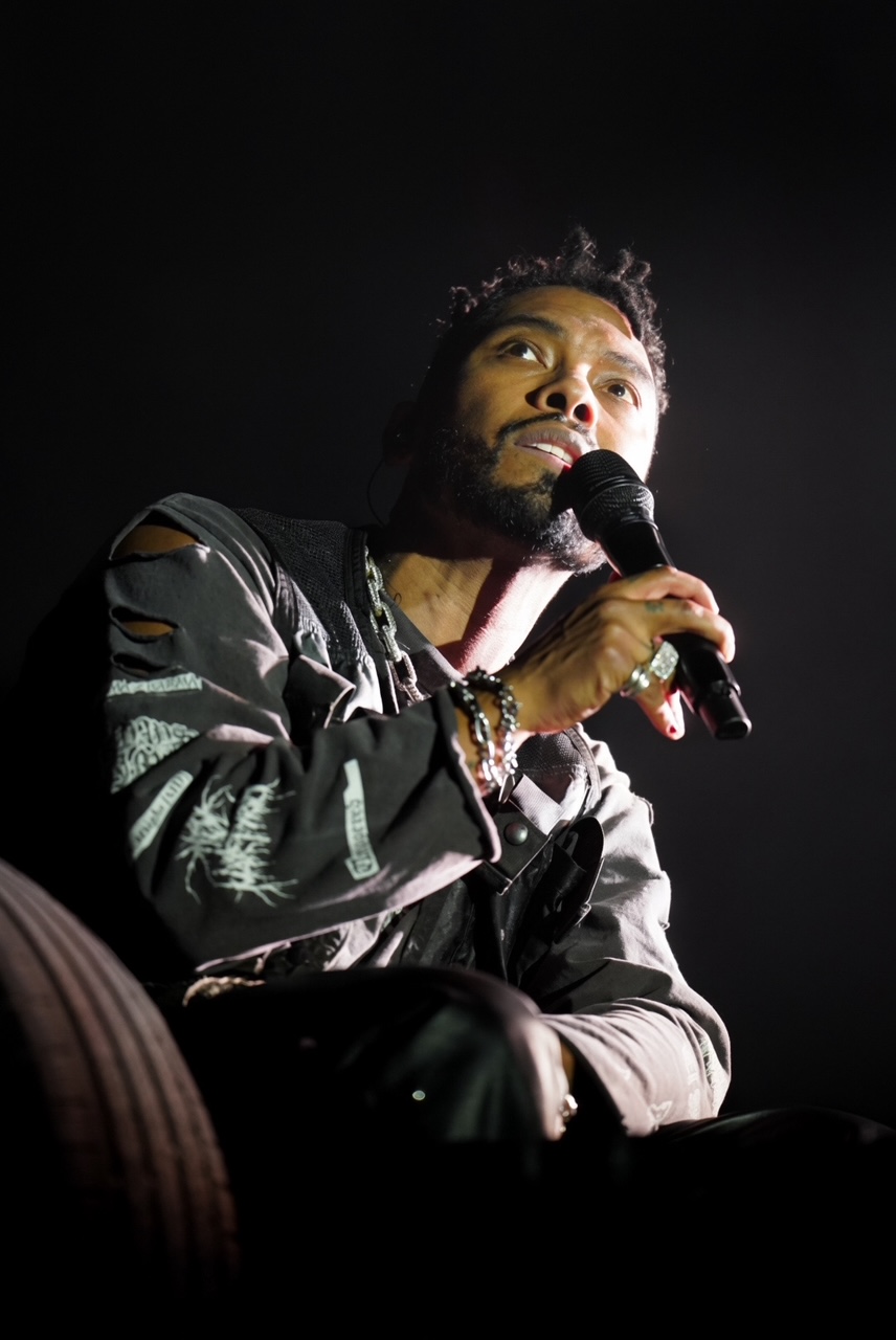 Singer Miguel lit by a single hard spotlight against a black stage, seated and singing into a handheld microphone, wearing a distressed grey jacket and layered chains