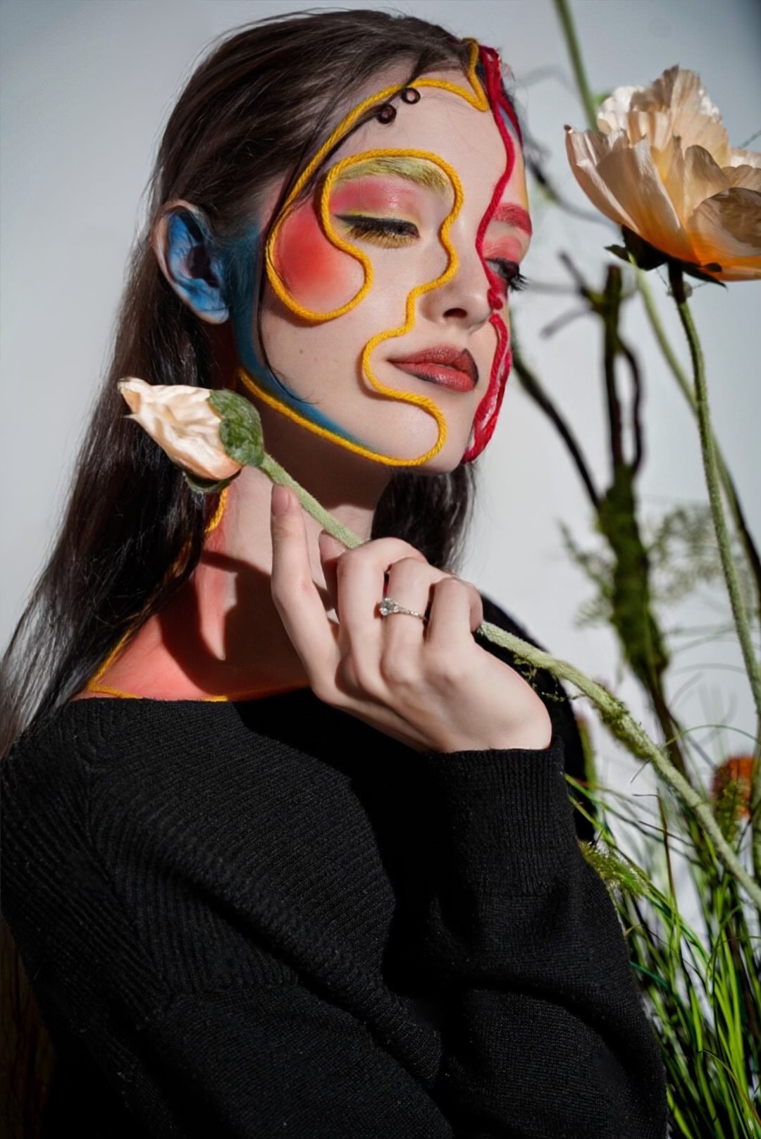 Editorial beauty portrait of a model whose face is traced with looping yellow, red and blue yarn over red and green eye makeup, holding a pale poppy against a grey backdrop