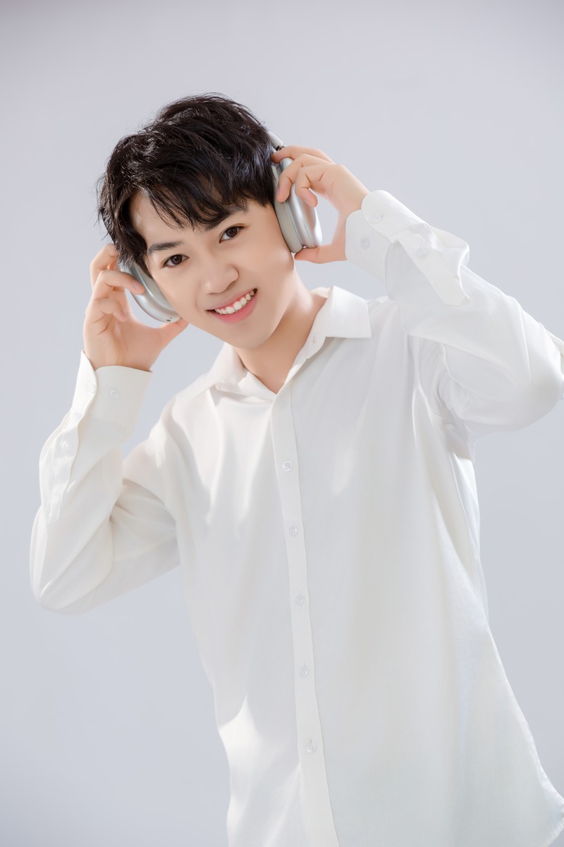 Composer Yuxuan Liu smiling in a white shirt, holding a pair of silver over-ear headphones to his ears against a light grey backdrop