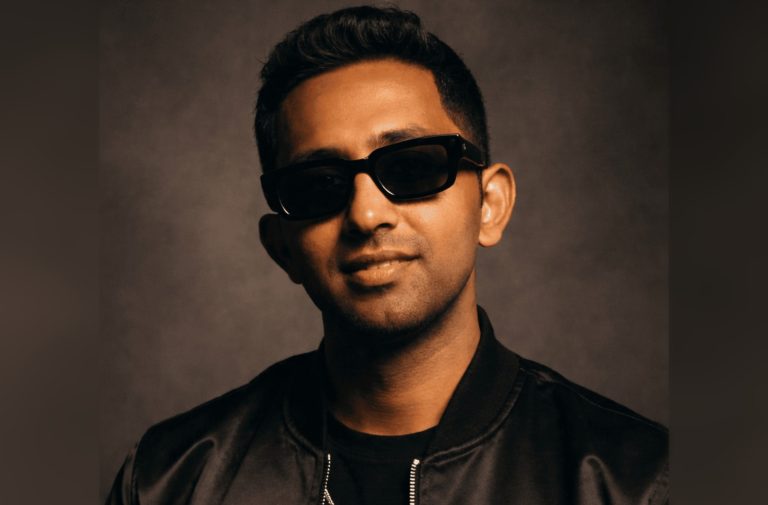 Australian R&B and Afrobeats artist Roger K in a black satin bomber jacket and dark rectangular sunglasses, photographed against a warm brown studio backdrop.