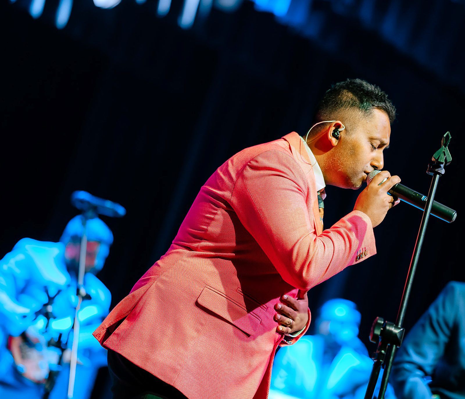 Roger K bent over a microphone stand mid-performance in a salmon-pink jacket, with a guitarist lit in blue behind him on a darkened stage.