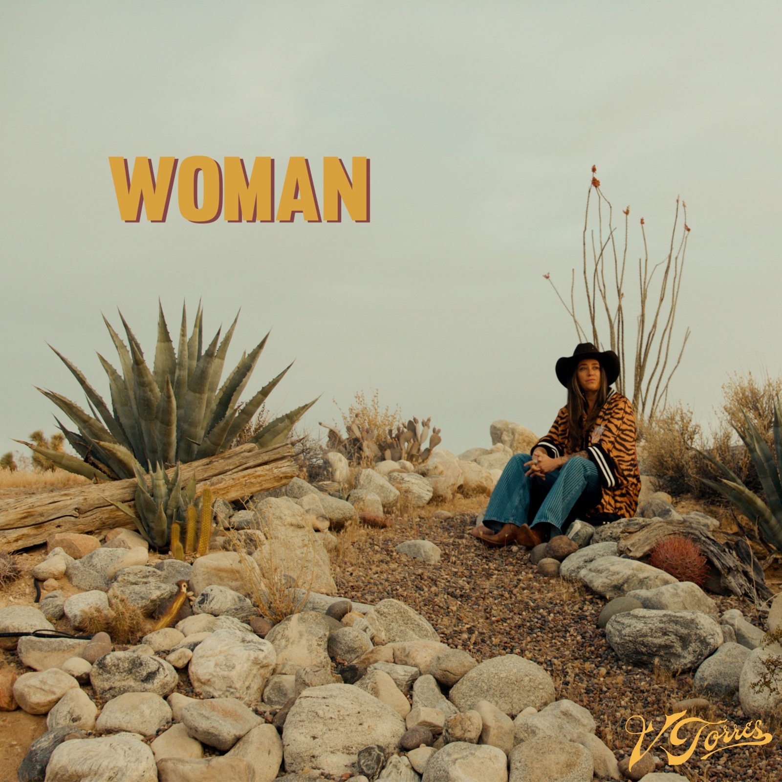 Single artwork for “Woman”: V Torres sits among rocks, agave and ocotillo in the high desert wearing a tiger-print coat and black hat, under the word WOMAN in gold.