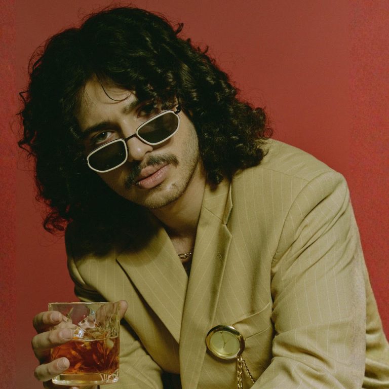 Official single artwork for “Lost & Never Found” by Davide Dalmonte: the singer in a pinstriped tan suit and white-framed sunglasses, holding a glass against a red backdrop.