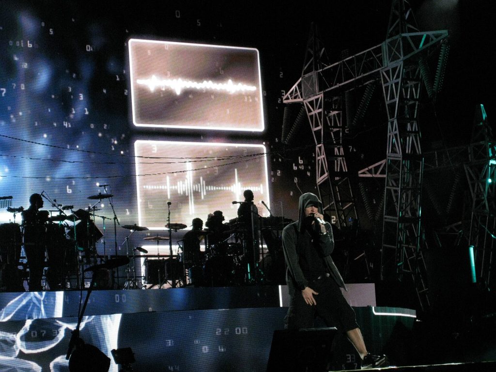 Eminem in a black hoodie performing on a large stage lit in blue with a band behind him