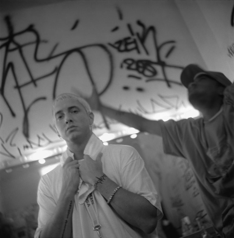 Black-and-white photo of a bleach-blond Eminem in a white T-shirt backstage in front of a graffiti wall