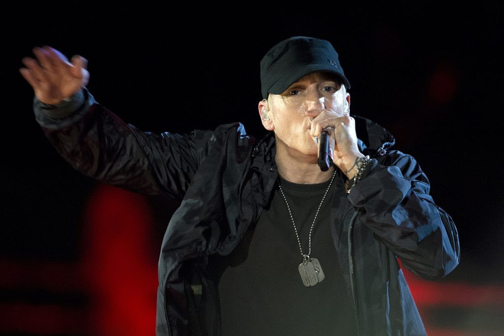 Eminem in a black cap and jacket rapping into a microphone with one arm extended