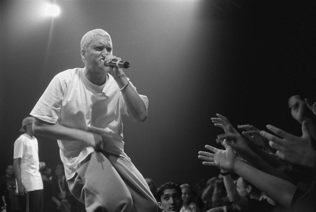 Black-and-white photo of a young, bleach-blond Eminem crouching at the edge of a stage as fans reach toward him