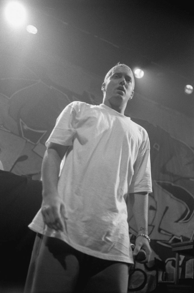 Full-length black-and-white photo of Eminem on stage in an oversized white T-shirt and baggy pants with bleached hair