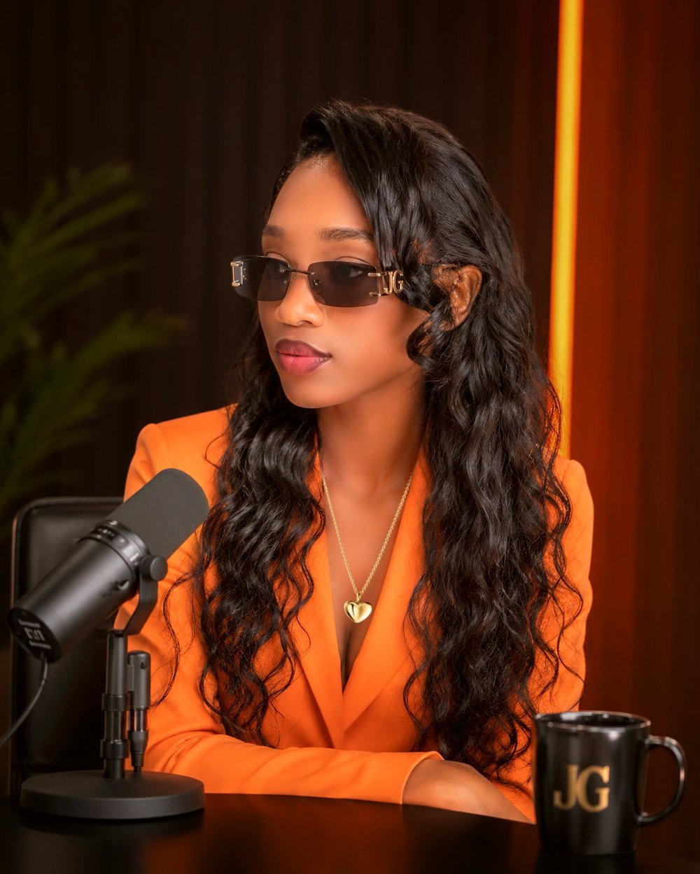 Jessygirl in an orange blazer and sunglasses seated at a podcast microphone beside a mug with her JG monogram