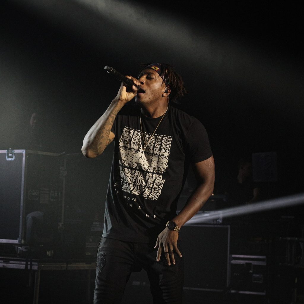 Lecrae performing on a dark stage, microphone raised, in a black graphic T-shirt