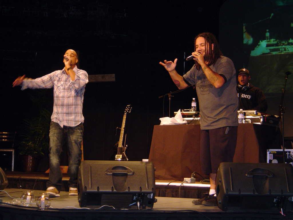 Pigeon John and RedCloud performing together on a small stage, each holding a microphone