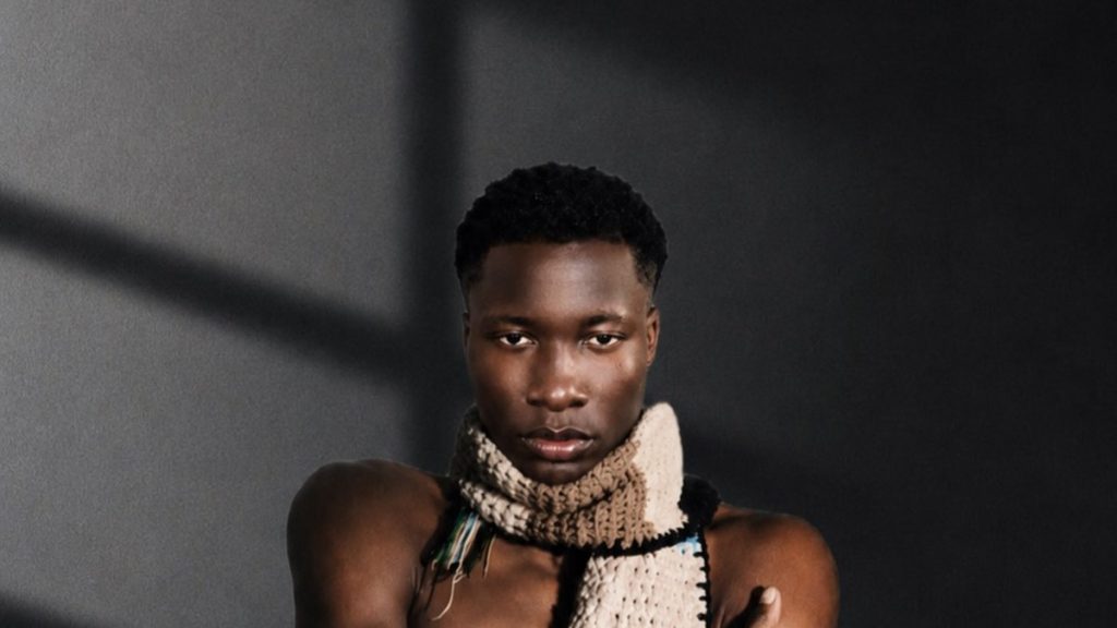 Trey Mavrik, Nigerian-born indie-pop artist, in a crocheted scarf with arms crossed against a grey wall