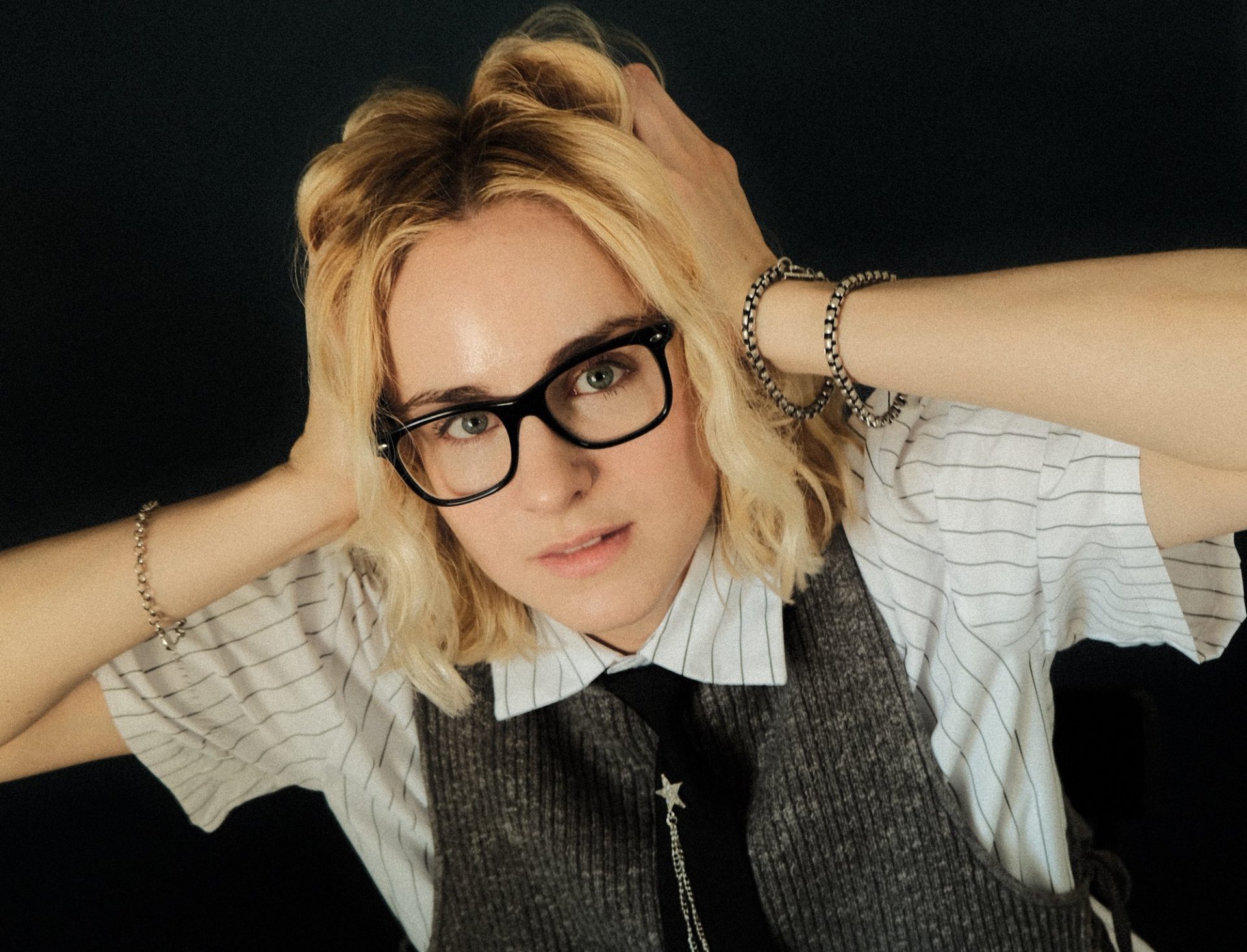 Analesa Fisher with hands in their hair, wearing glasses, a striped shirt and a gray knit vest