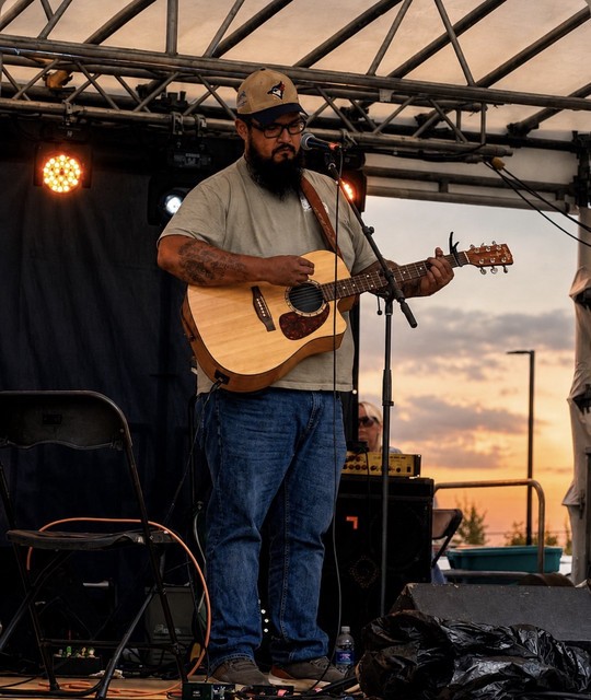 B Fabian playing acoustic guitar on an outdoor stage at sunset