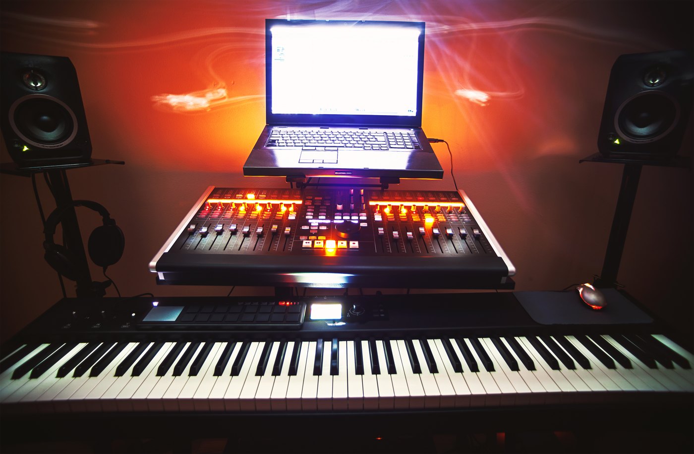 A music production desk with a MIDI keyboard, mixing controller and laptop between two studio monitors