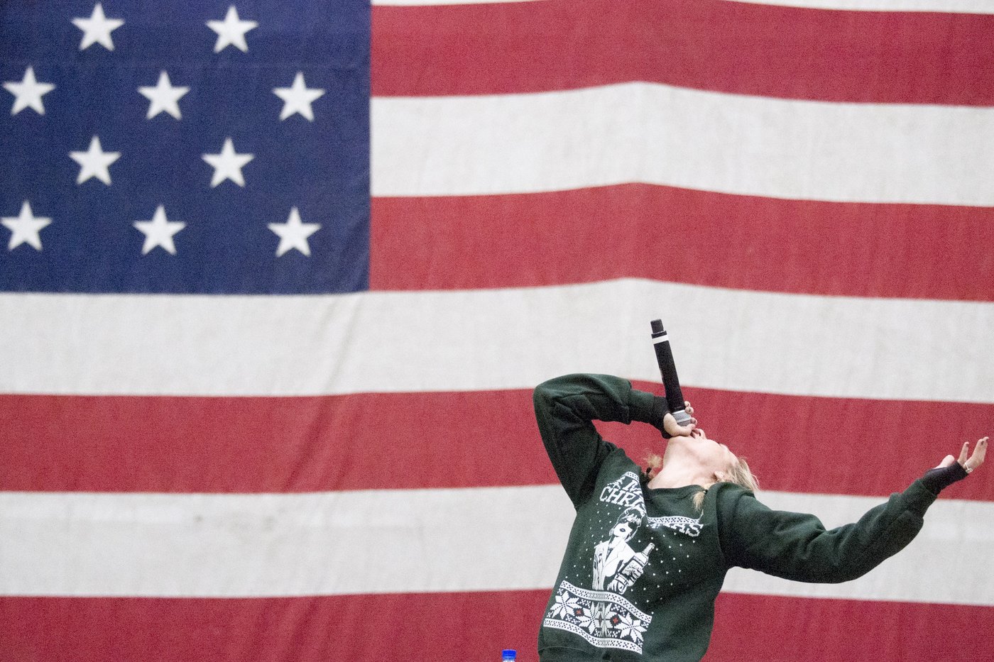 Iliza Shlesinger leaning back with a microphone in front of a giant American flag at Bagram Airfield, Afghanistan, on Christmas Eve 2017