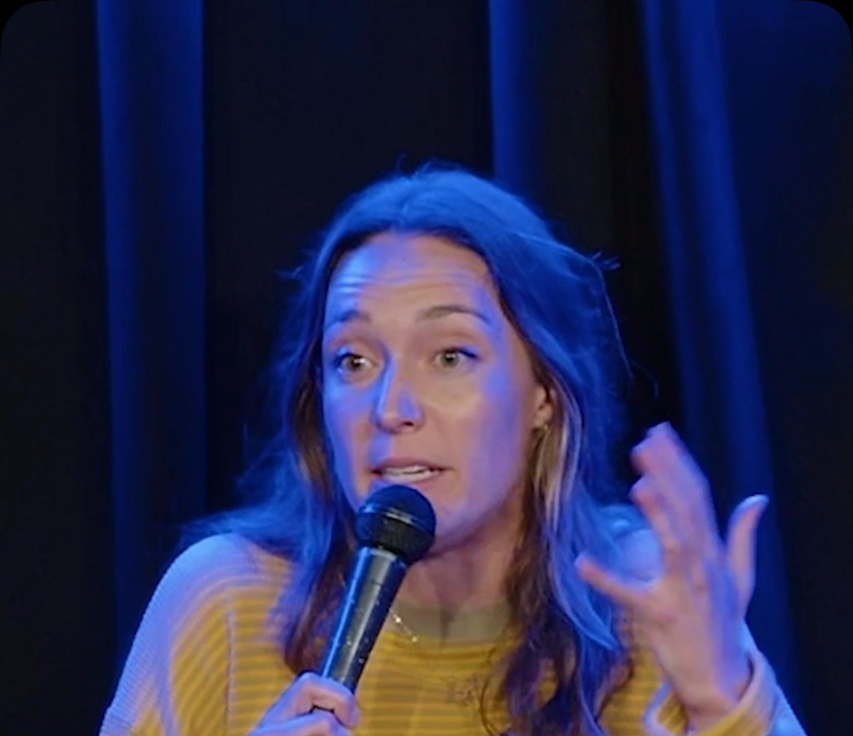 Kat Bird performing stand-up comedy on stage holding a microphone