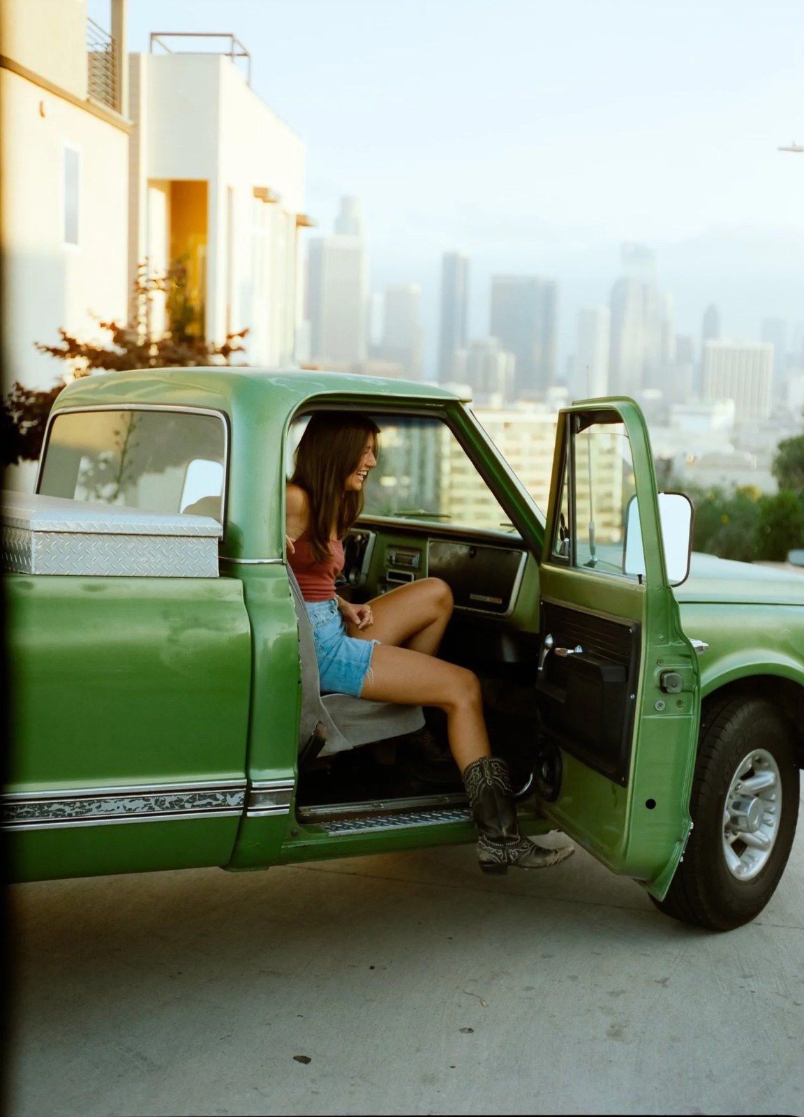 Vienna Hintze sitting in the driver's seat of Fiona, her green vintage pickup, with the downtown Los Angeles skyline behind her
