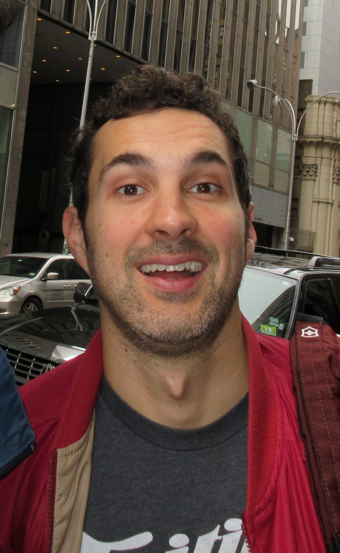Comedian Mark Normand smiling on a Manhattan sidewalk in a red jacket, March 2018