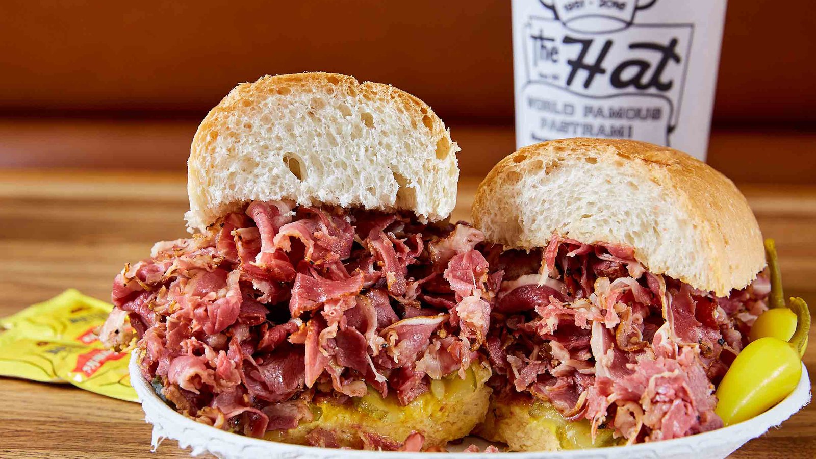 A pastrami sandwich piled high on French rolls with pepperoncini in front of a cup from The Hat