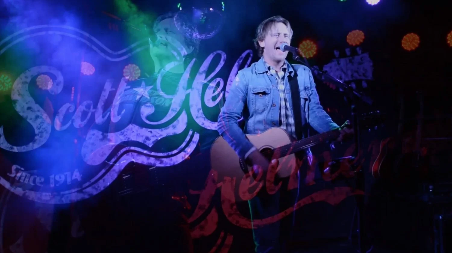 Scott Helmer performing live on stage with an acoustic guitar